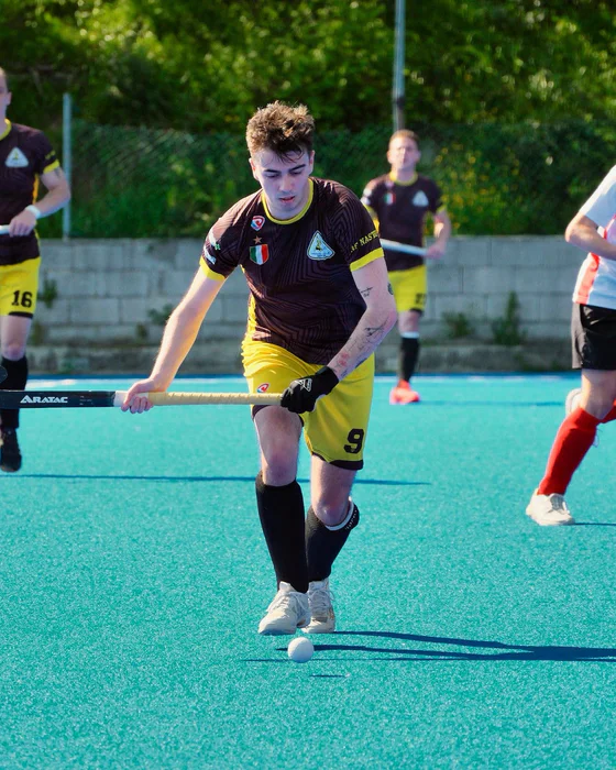 Andrea Bertorello running with the ball on a blue hockey pitch.